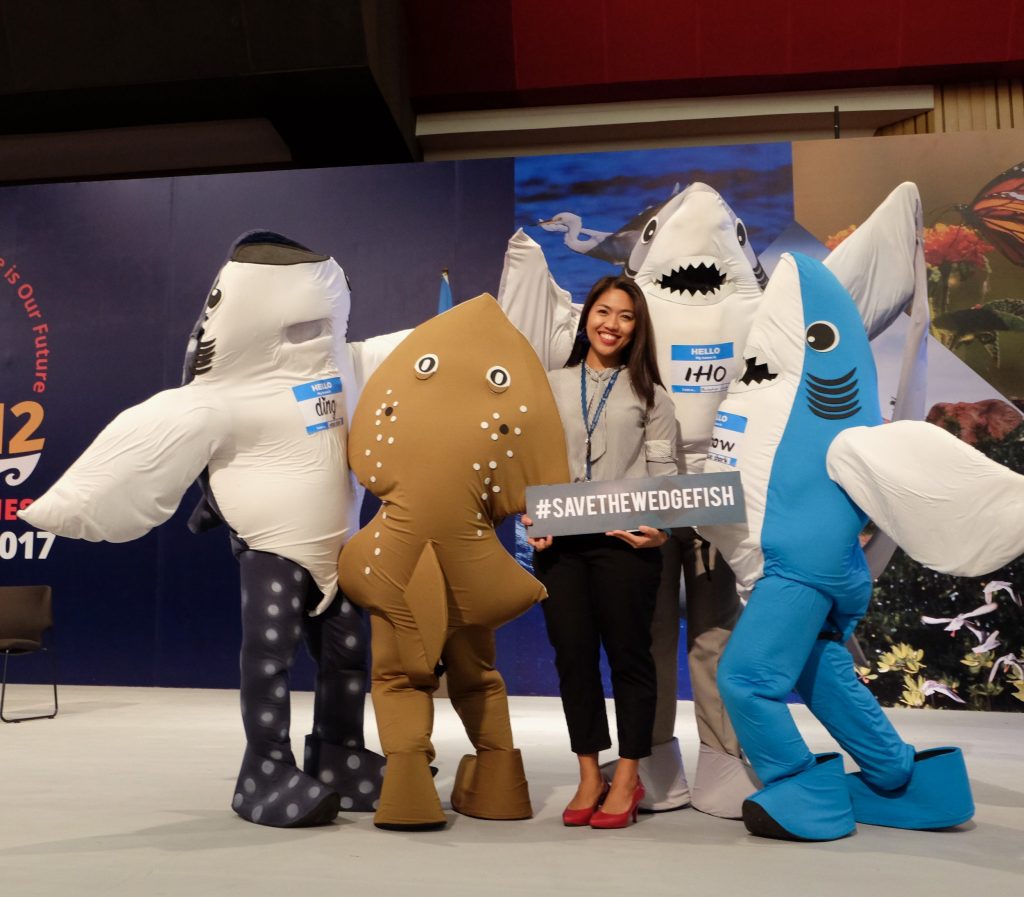 Ana Oposa holding a sign saying #savethewedgefish surrounded by people wearing fish costumes including a whale shark, stingray and two other sharks.