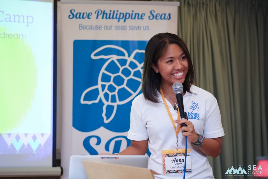 Ana Oposa holding a microphone and presenting to an unseen audience with a Save Philippine Seas banner in the background.