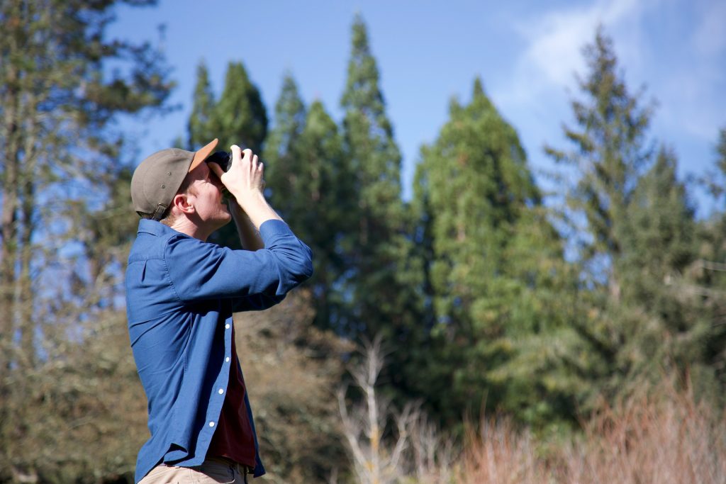 Charlie, dressed in a long-sleeve blue shirt, stands in a forested area holding a camera and taking a photo of something outside of the shot.