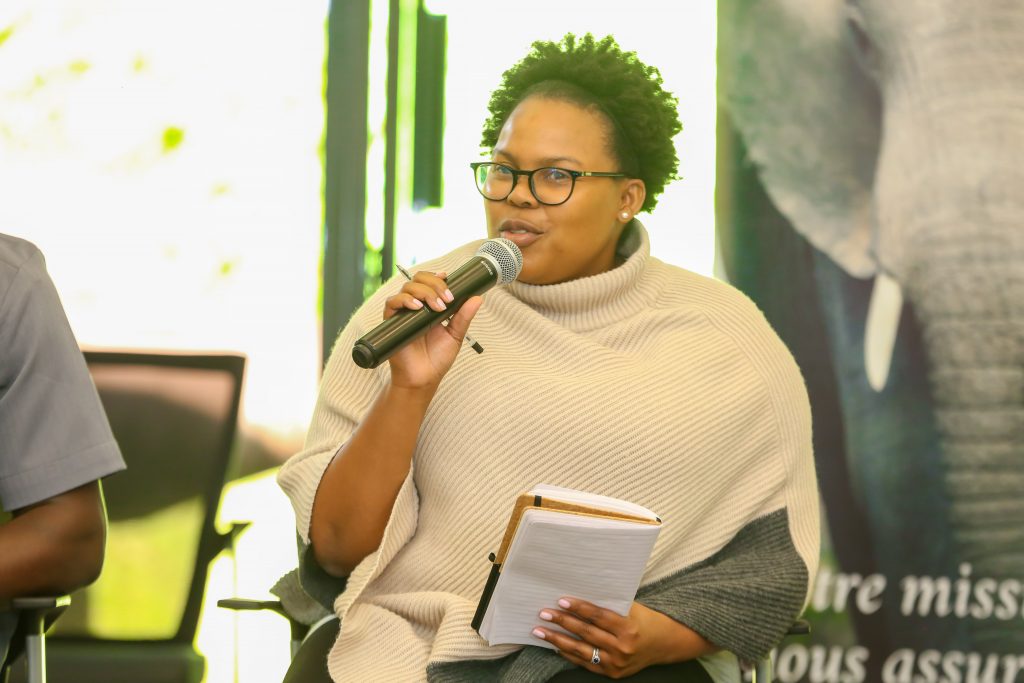 Simangele Msweli-Ratsoana sitting on a stool holding a microphone and notepad presenting to an unseen audience.