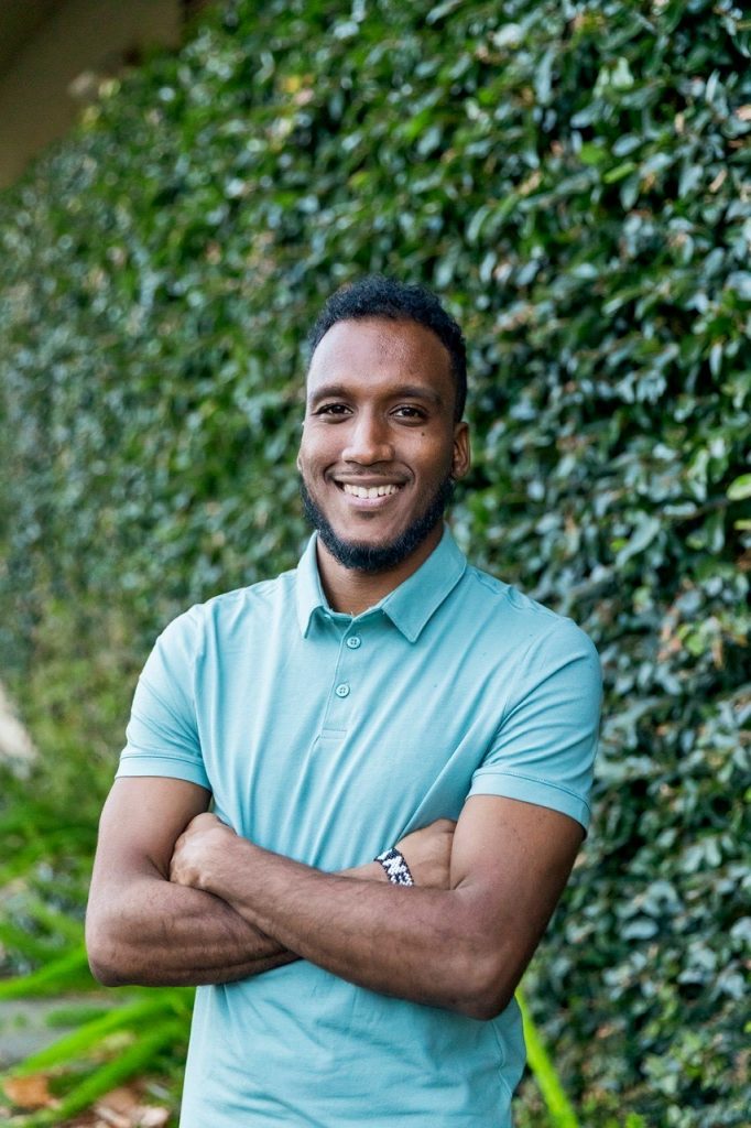 A head and shoulders profile photo of Joshua Prentice standing in front of a wall covered in leaves