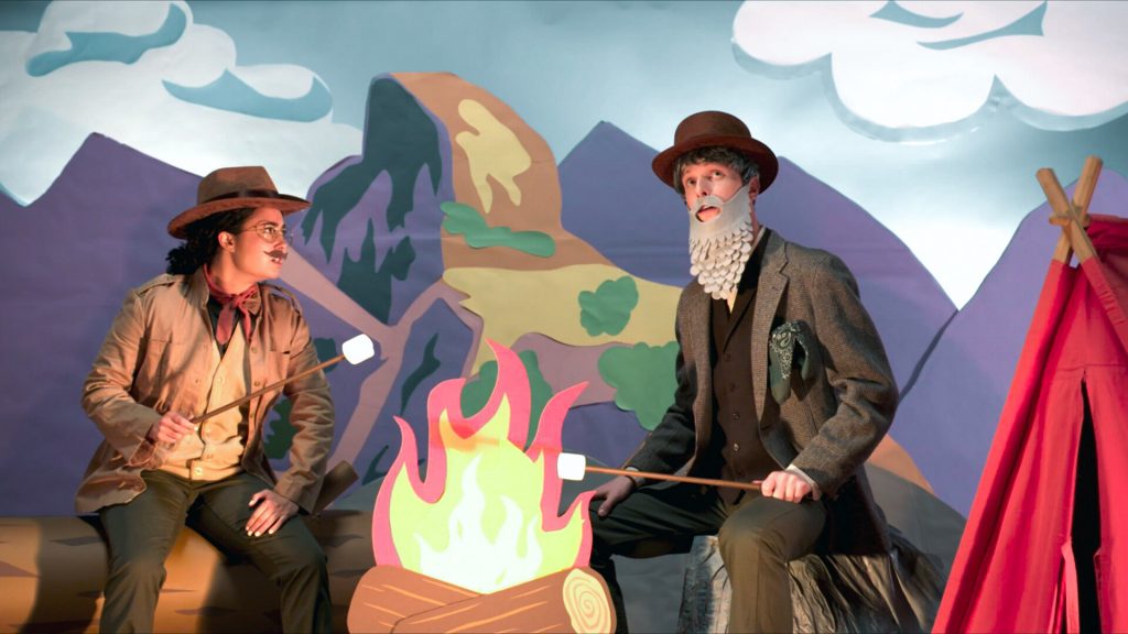 An image from Charlie and a colleague dressed up on the set of Weird but True sitting either side of a decorative cutout campfire.