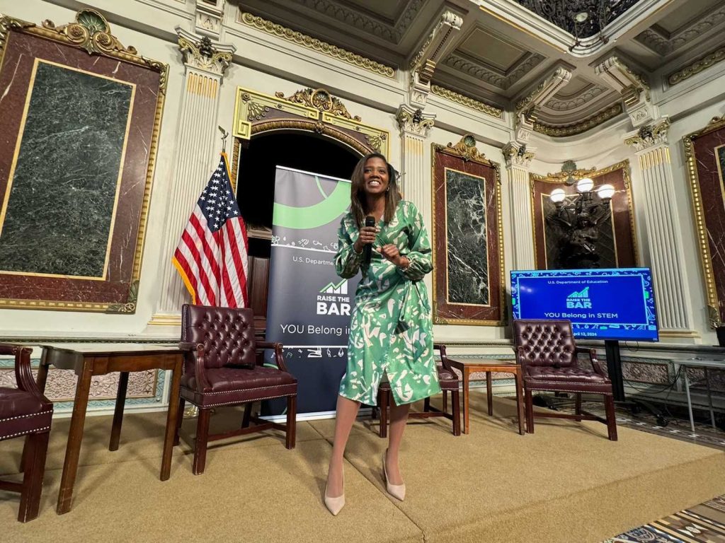 Yamille Toussaint stands in front of a pull up banner reading ' Raise the BAR' at a room in the White House