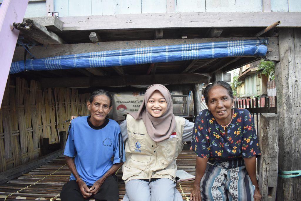 Nuha, smiling, sits with her arm around two other women. On her left fisherwoman Khadija dressed in loose pants and a loose blue t-shirt and Khadasiah dressed in a colourful dress and t-shirt with yellow, red and blue flowers printed on it.