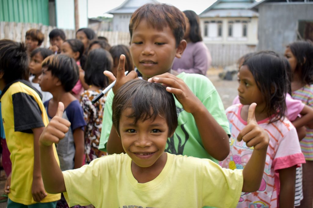 A small child in a yellow t-shirt stands smiling giving two thumbs up to the camera while a taller, older child stands behind him resting a hand on his head while holding a marker in his other hand, as if he's about to write on the T-shirt of the child in front of him.