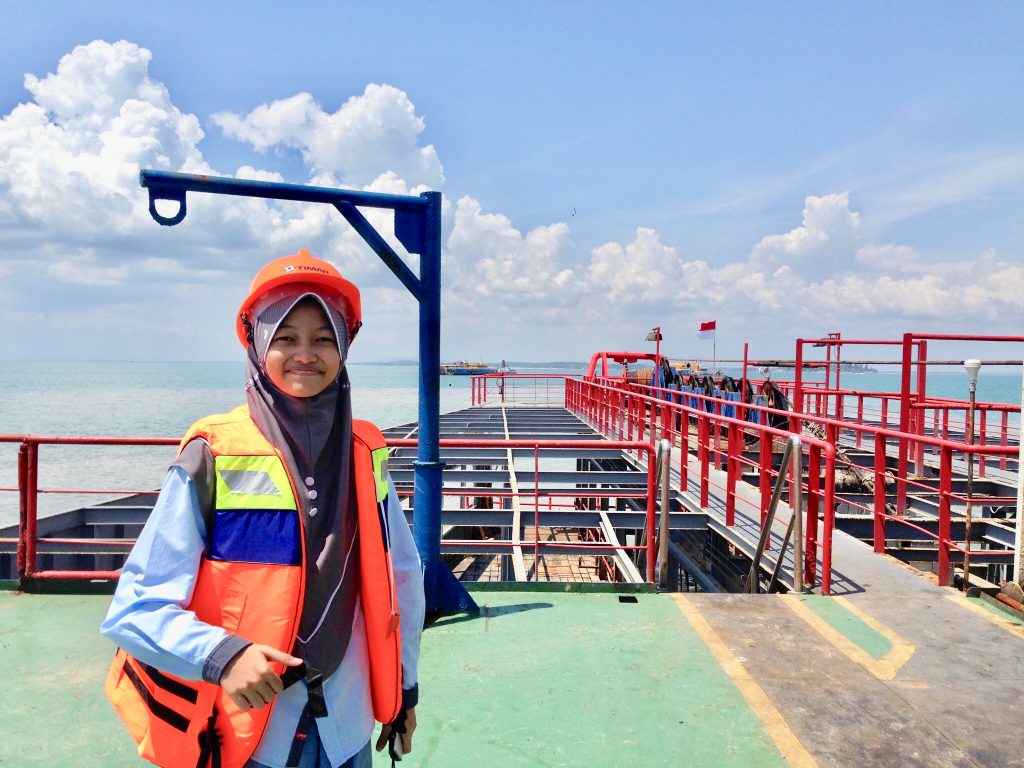 16-year-old Nuha Anfaresi wears a long-sleeve blue shirt, life jacket and orange construction helmet on a suction dredge site with a calm ocean in the background